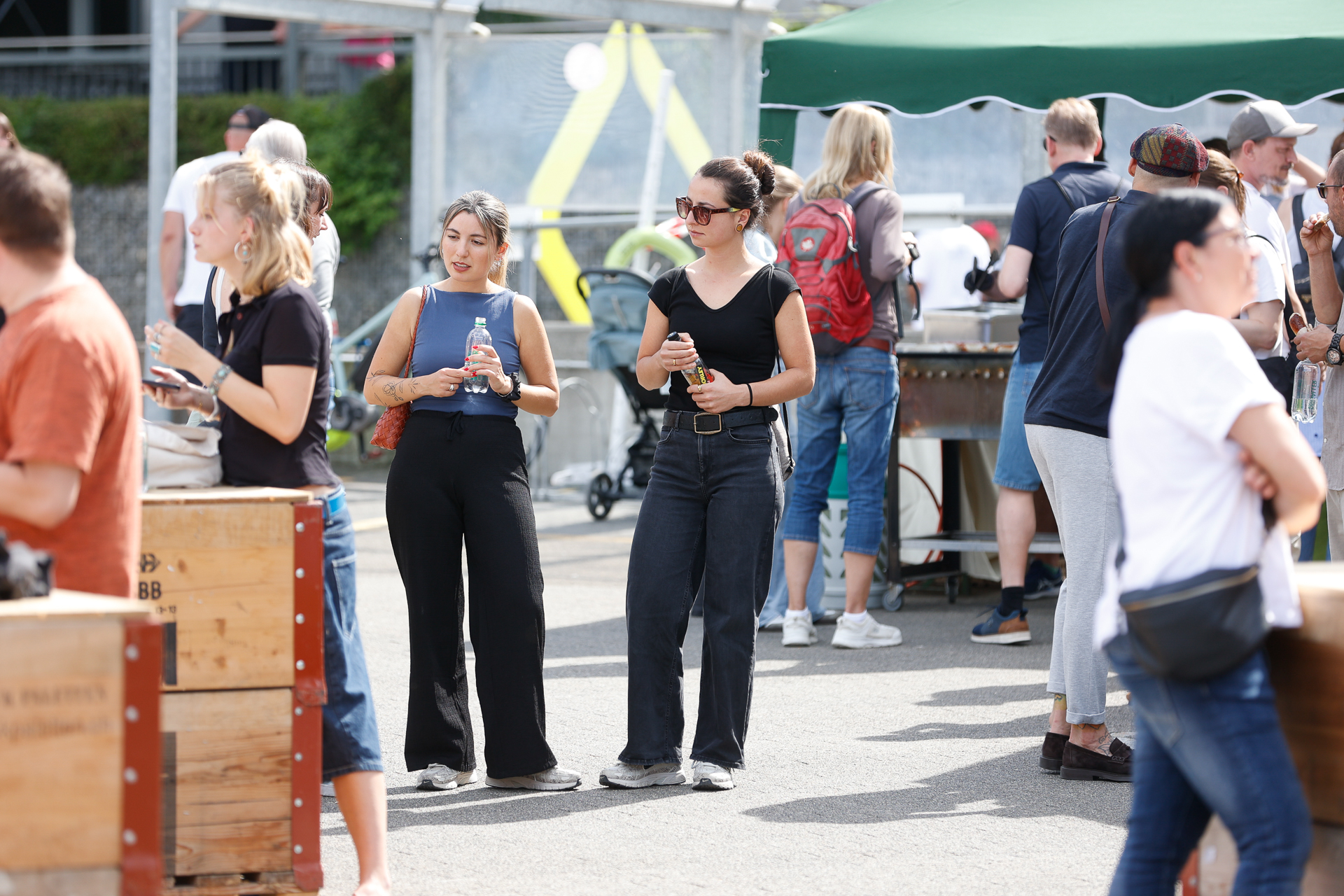 Eine Gruppe von Menschen versammelt sich im Freien bei einer Veranstaltung; einige halten Getränke in der Hand und unterhalten sich, im Hintergrund sind Imbissstände und Holzkisten zu sehen.