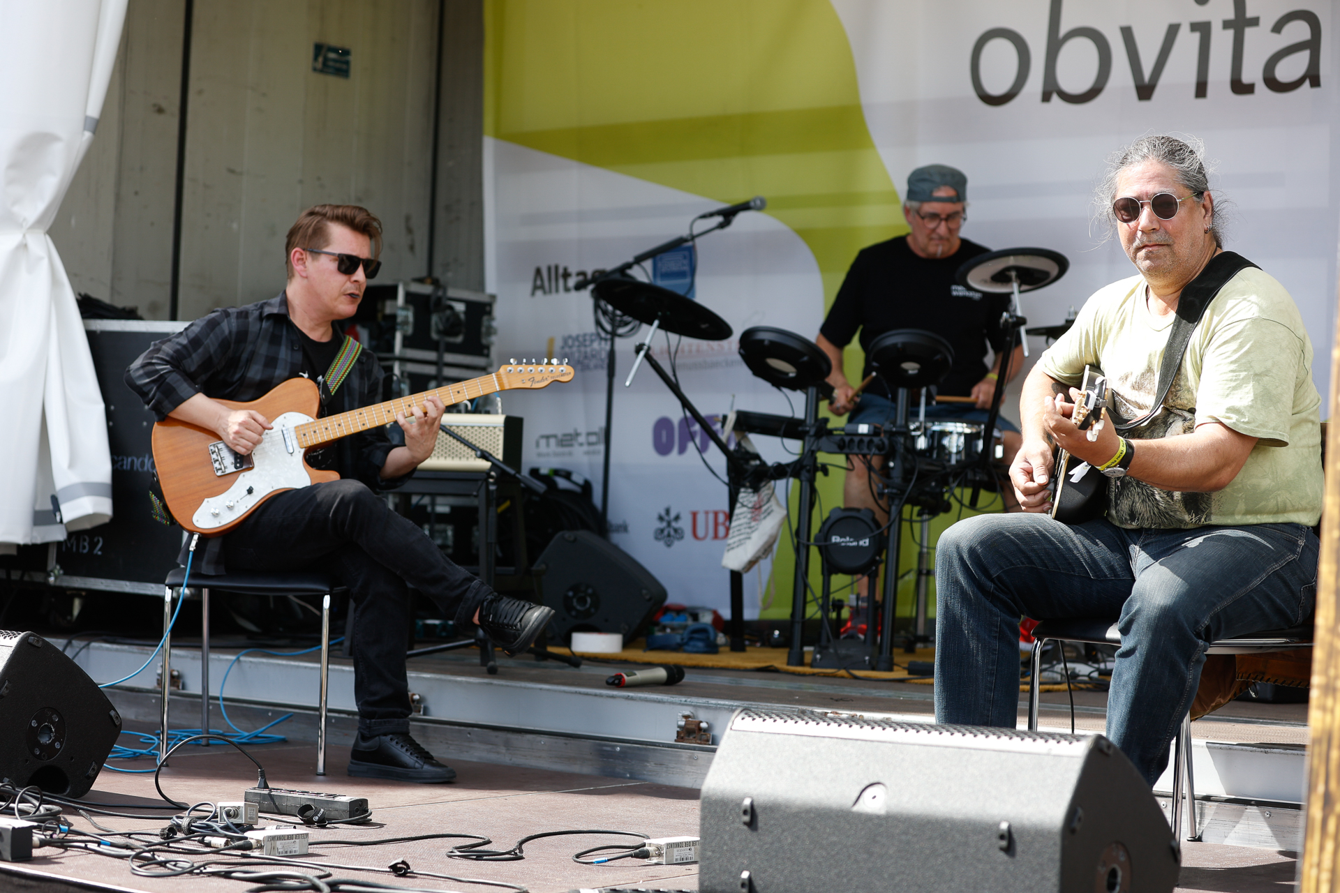 Zwei Gitarristen sitzen da und spielen E-Gitarre, während hinter ihnen ein Schlagzeuger auf einer Freiluftbühne mit Mikrofonen, Lautsprechern und einem Banner im Hintergrund, auf dem „obvita.“ steht, ein elektronisches Schlagzeug spielt.