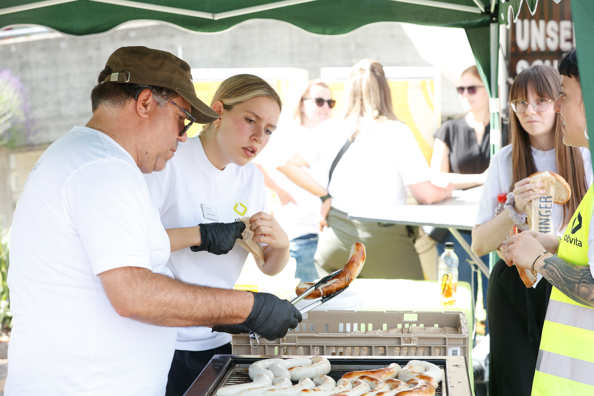 Bei einer Veranstaltung im Freien stehen zwei Personen unter einem Zelt und grillen gemeinsam Würstchen, während im Hintergrund andere in weißen Hemden miteinander reden und essen. Eine Person wendet die Würstchen mit einer Zange auf dem Grill.