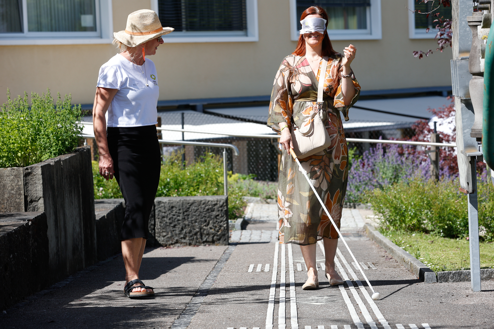 Eine Frau mit einer Augenbinde und einem weißen Blindenstock geht auf einem taktilen Pfad entlang, während eine andere Frau mit Hut und weißem Hemd neben ihr im Freien hergeht und sie offenbar führt oder beobachtet.