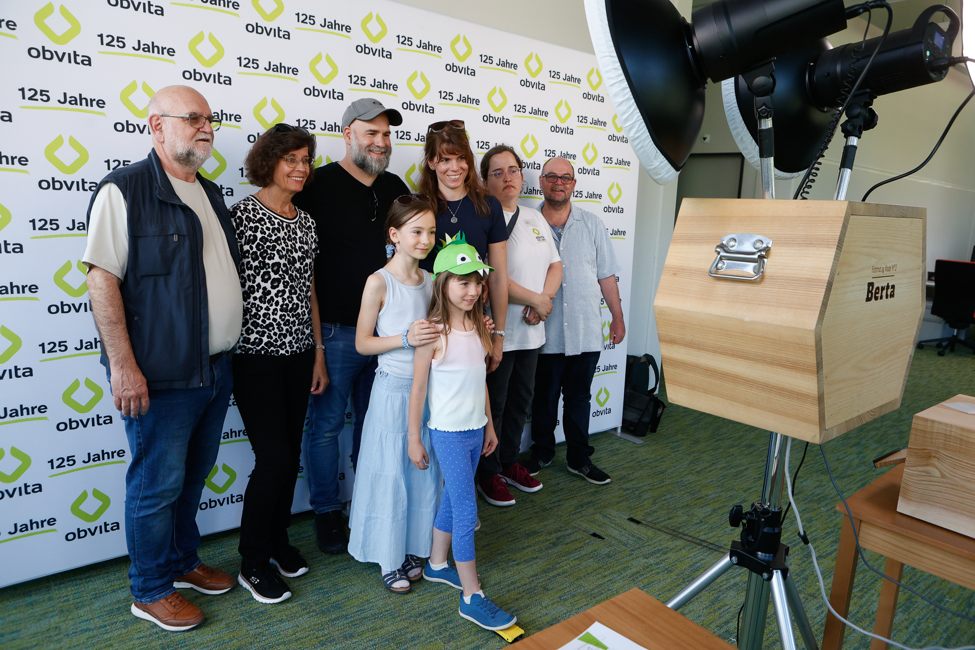 Eine Gruppe aus sieben Erwachsenen und zwei Kindern posiert für ein Foto vor einem Step-and-Repeat-Banner mit den Aufschriften „125 Jahre“ und „obvita“. Rechts ist eine große Kameraausrüstung zu sehen.