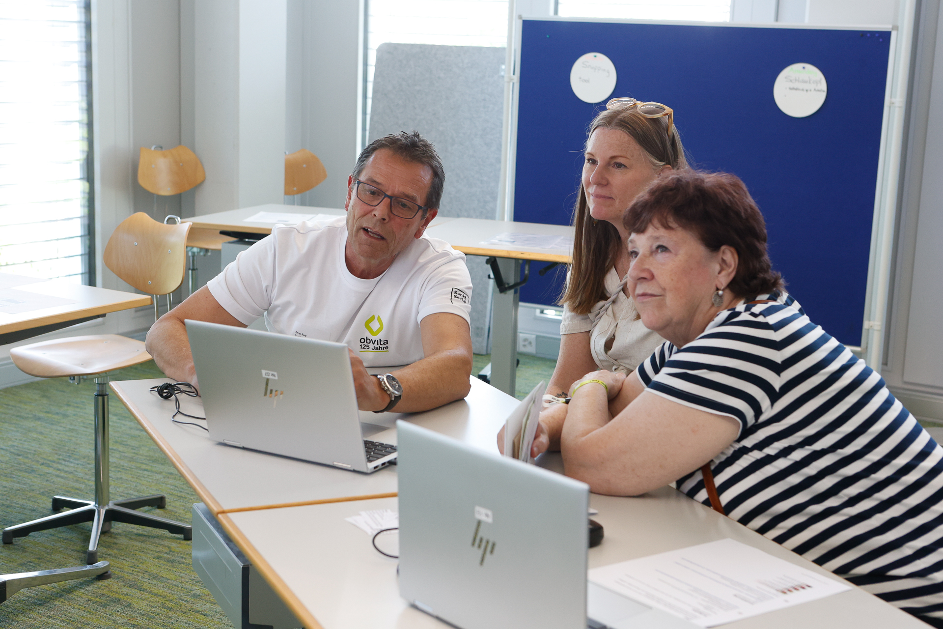 Drei Erwachsene sitzen an einem Tisch in einem hellen Klassenzimmer und arbeiten gemeinsam an ihren Laptops. Zwei Frauen und ein Mann blicken nach vorne und scheinen sich zu unterhalten oder einer Präsentation zu folgen.