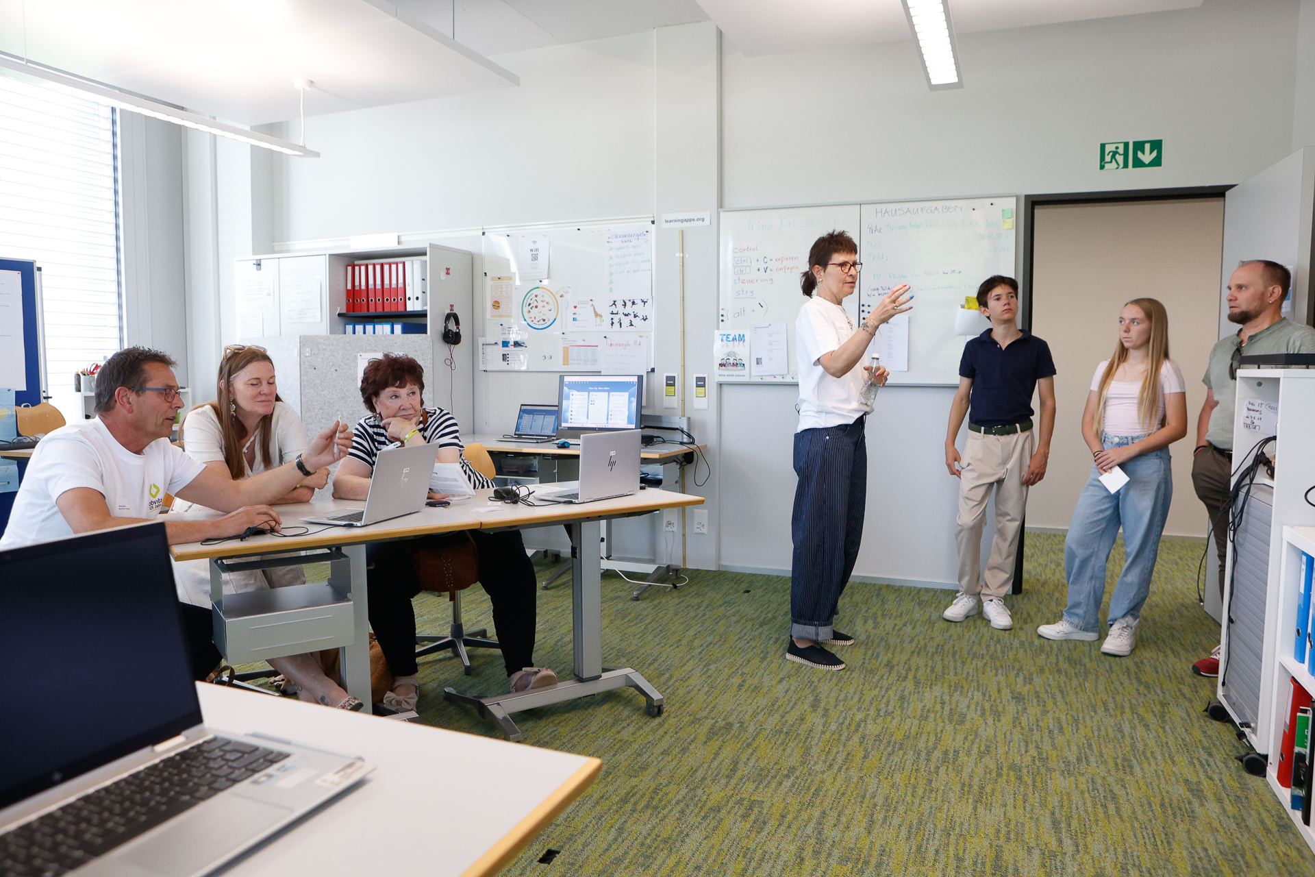 In einem modernen Büro-Unterrichtsraum befinden sich sechs Personen: Drei sitzen an Schreibtischen mit Laptops, während drei andere in der Nähe einer Weißwandtafel stehen. Eine der stehenden Personen gestikuliert beim Sprechen. Der Raum ist hell und verfügt über weiße Wände, Fenster und Computerausstattung.