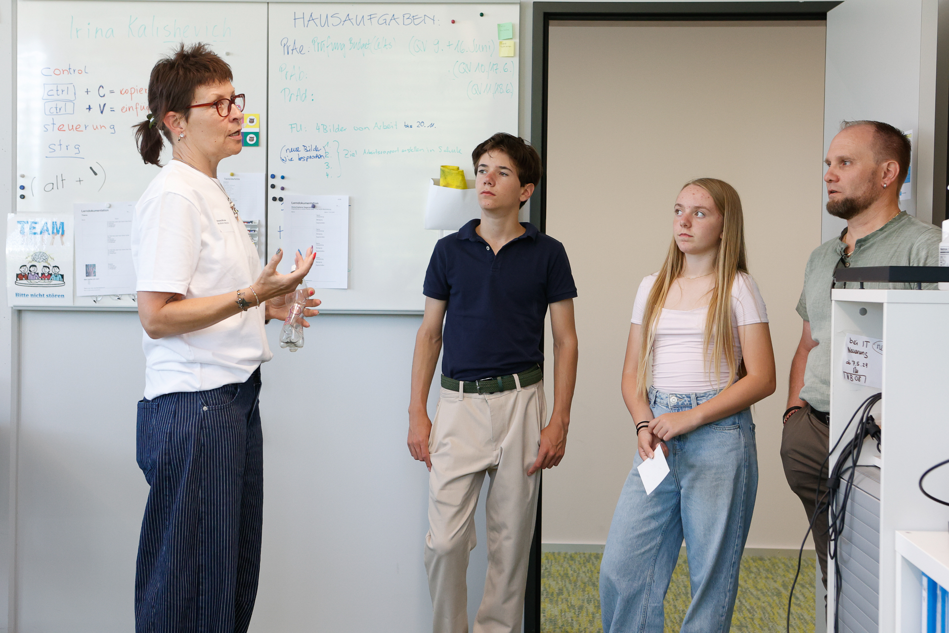 Eine Frau unterhält sich mit drei Personen – einem Mann, einem Teenager und einem Mädchen –, die in einem Büro vor einer Whiteboard stehen, auf der Notizen und Termine vermerkt sind. Alle wirken aufmerksam und sind in das Gespräch vertieft.