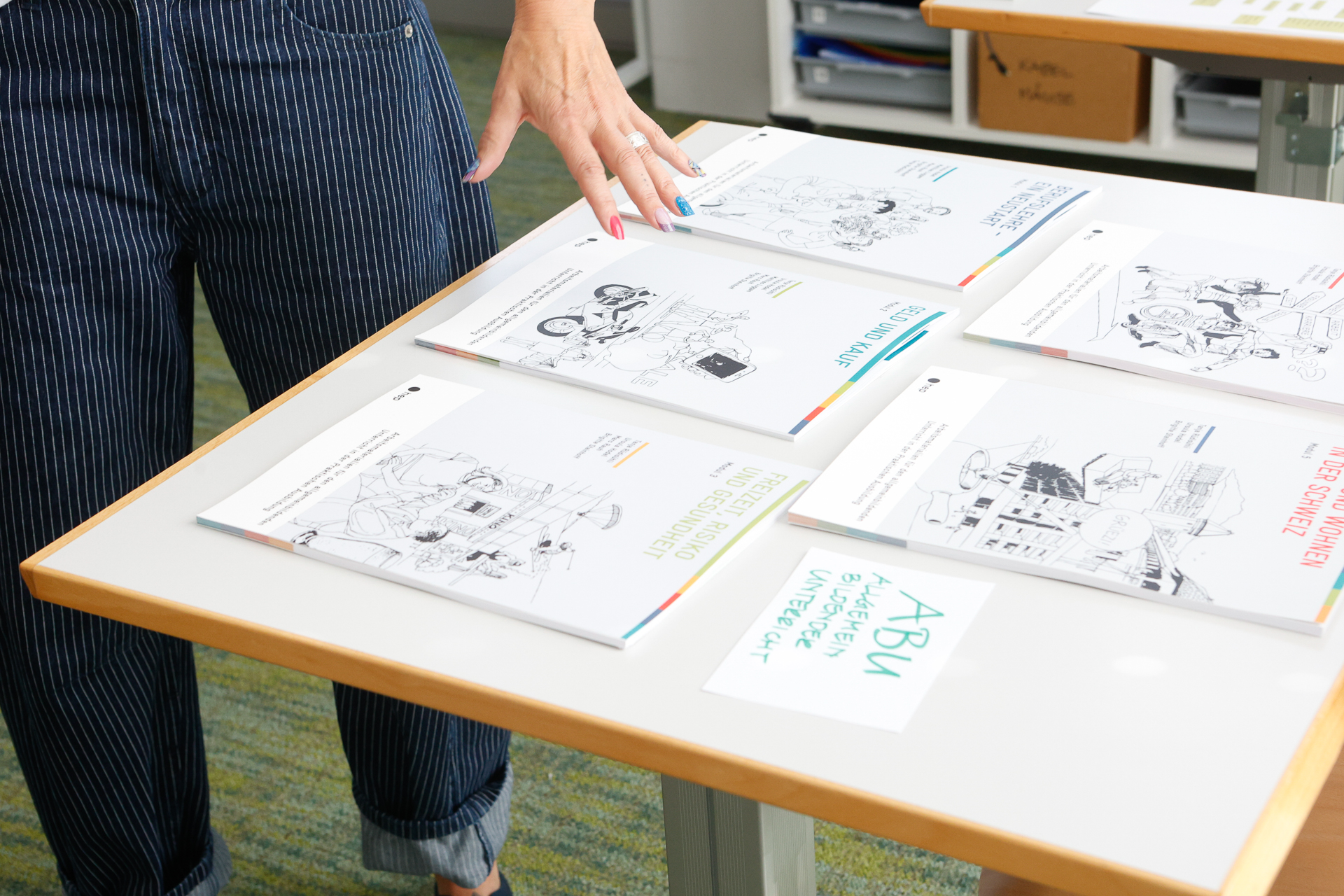 In einem hell beleuchteten Raum mit Büromöbeln steht eine Person mit lackierten Fingernägeln neben einem Tisch, auf dem mehrere illustrierte Broschüren und ein handgeschriebenes Schild mit der Aufschrift „ABU“ ausliegen.