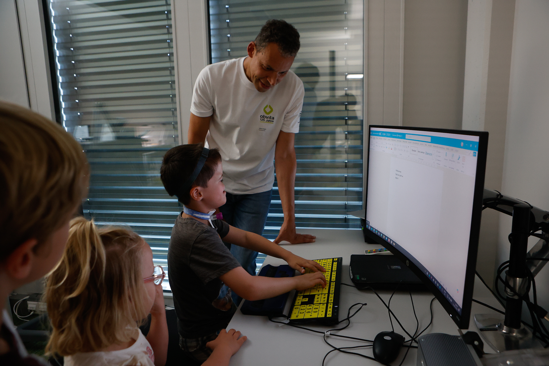 Ein Mann hilft einem kleinen Jungen, der an einem Computer eine Tastatur mit großer Schrift und hohem Kontrast benutzt. Zwei weitere Kinder schauen in einem hellen Raum mit großen Fenstern und Jalousien von nebenan zu.