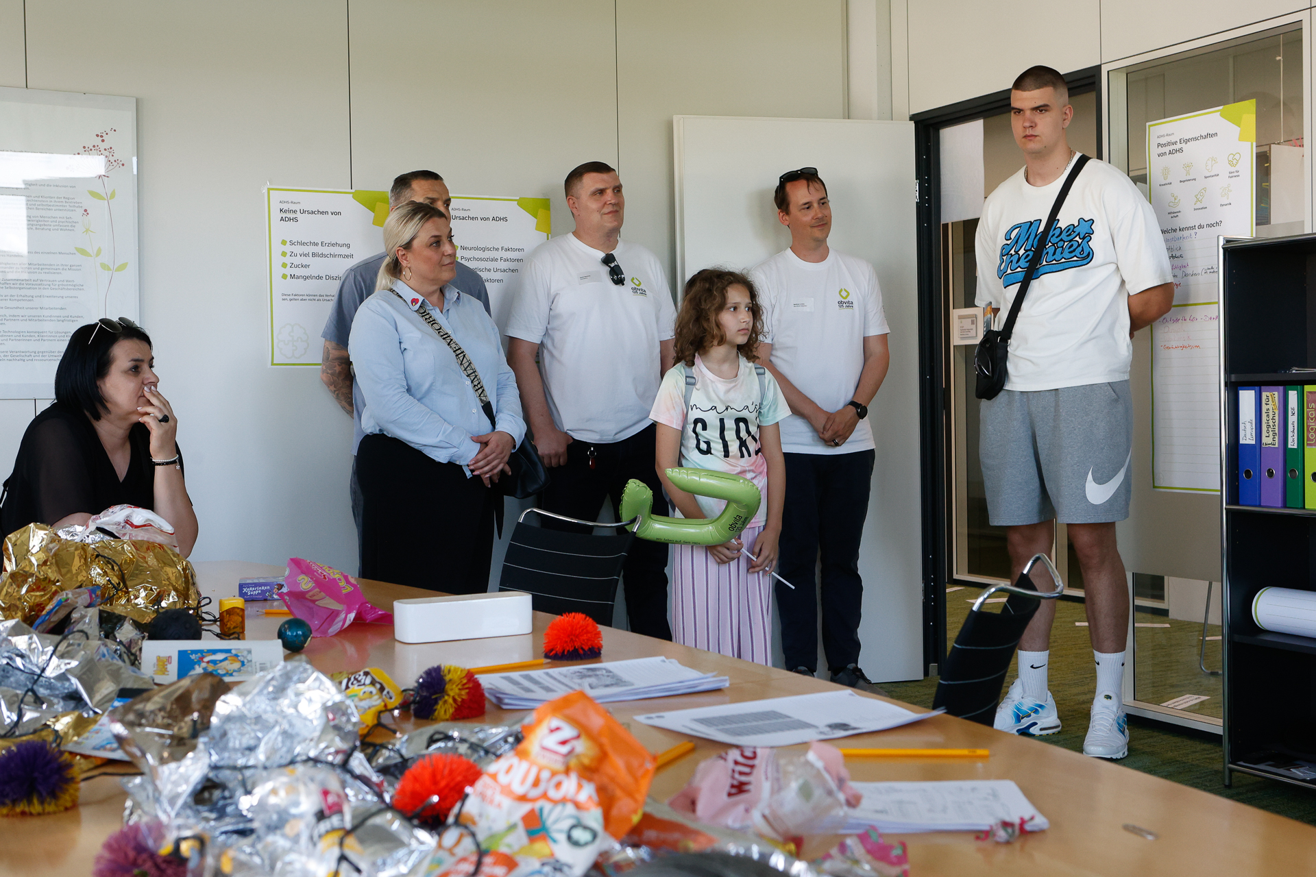 Eine Gruppe von Erwachsenen und ein Kind stehen in einem Raum in der Nähe einer Tür und beobachten aufmerksam etwas. Im Vordergrund steht ein Tisch, der mit buntem Bastelmaterial und Papier bedeckt ist.