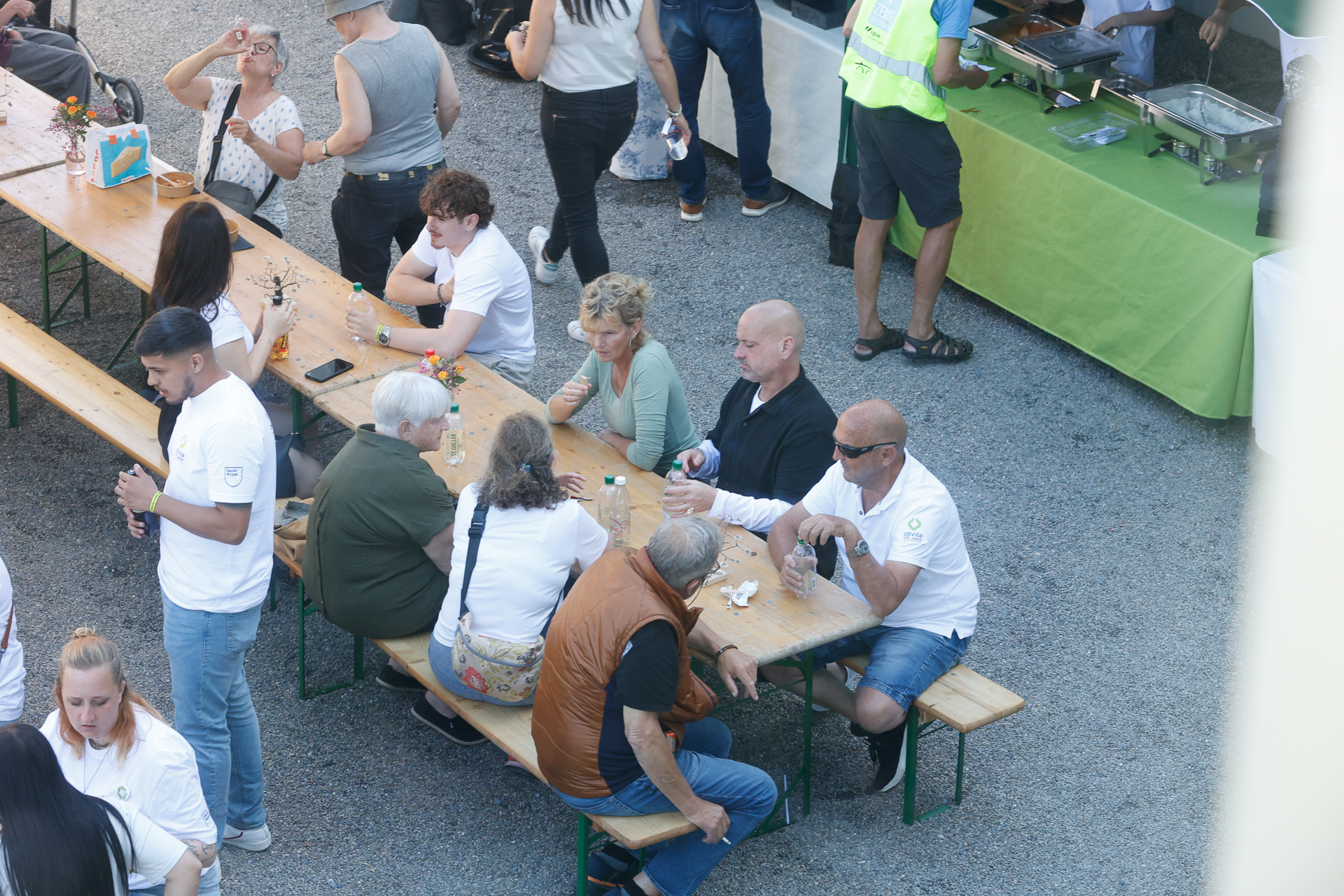 Eine Gruppe von Menschen sitzt und steht im Freien um lange Holztische herum; einige essen und trinken. Im Hintergrund ist eine Essensausgabe mit grünen Tischdecken zu sehen, und die Szene scheint bei einer ungezwungenen Veranstaltung oder einem Treffen im Freien stattzufinden.