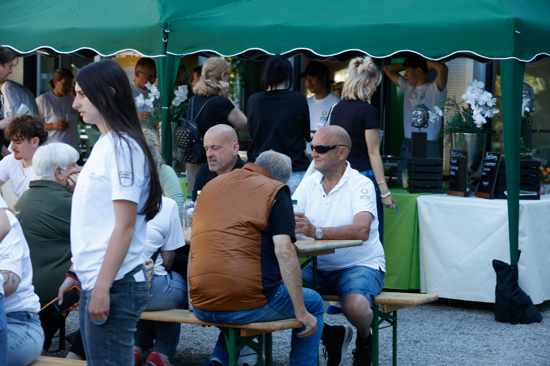 Unter einem grünen Zelt haben sich Menschen an Tischen im Freien versammelt; einige sitzen und unterhalten sich, andere stehen in der Nähe einer Theke. Die meisten sind leger gekleidet, und es scheint sich um eine gesellschaftliche oder gemeinschaftliche Veranstaltung zu handeln.
