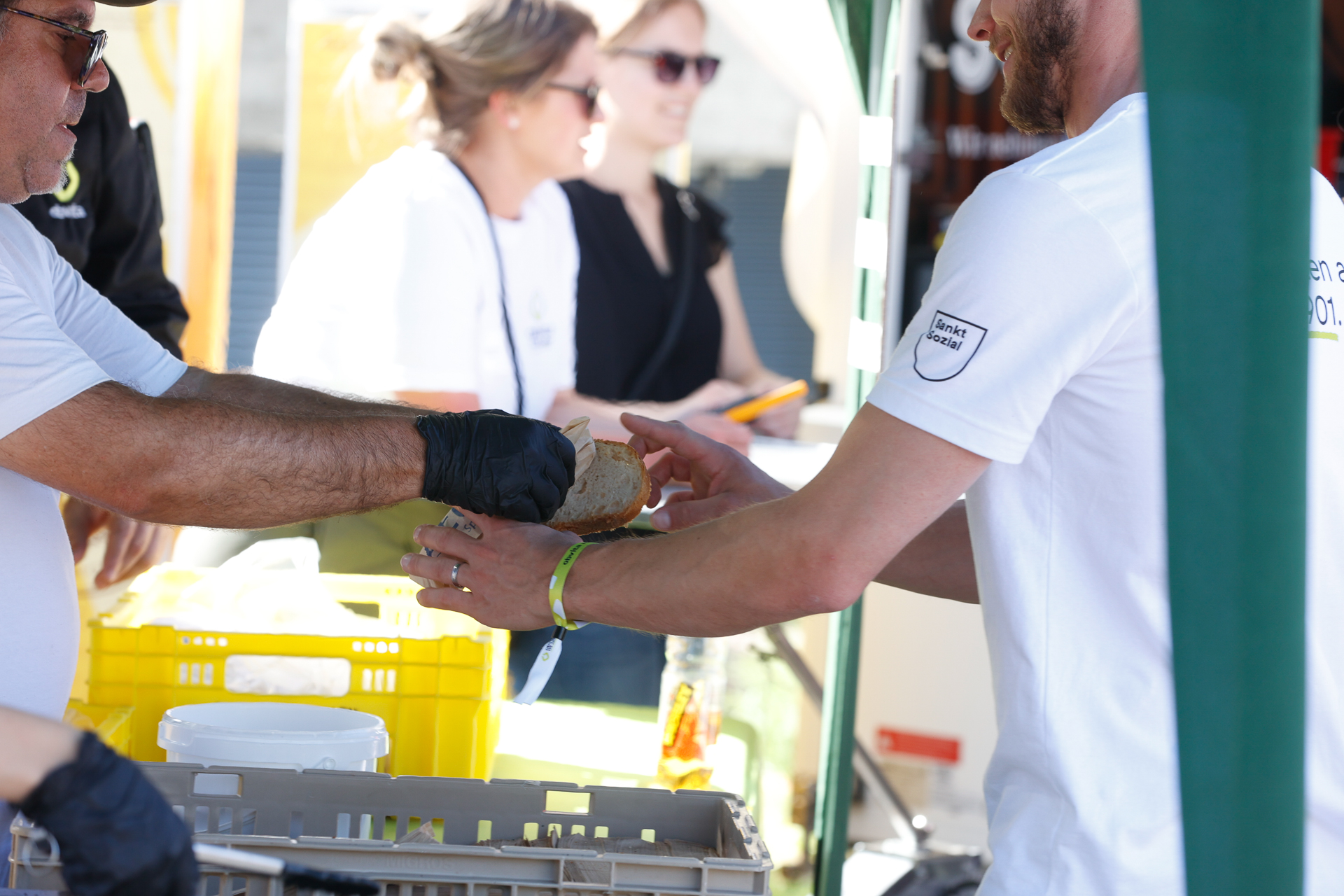 Zwei Personen, die beide weiße Hemden und schwarze Handschuhe tragen, tauschen an einem Imbissstand im Freien ein Sandwich aus, während im unscharfen Hintergrund zwei Frauen stehen und sich unterhalten.