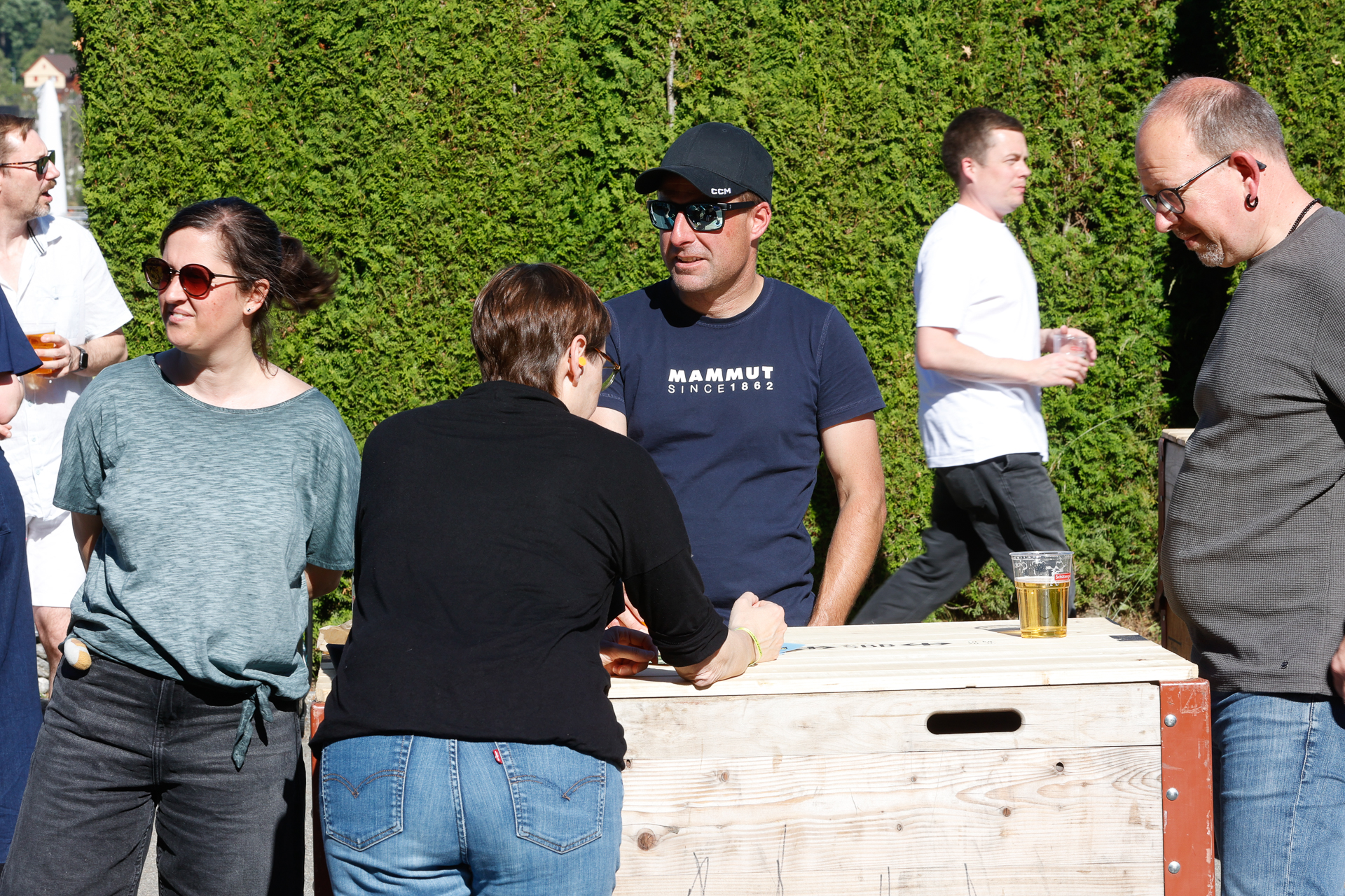 An einem sonnigen Tag stehen vier Personen im Freien um einen Holztisch herum und unterhalten sich. Eine Person hat ein Getränk auf dem Tisch stehen. Im Hintergrund geht ein Mann in einem weißen Hemd an einer hohen grünen Hecke vorbei.