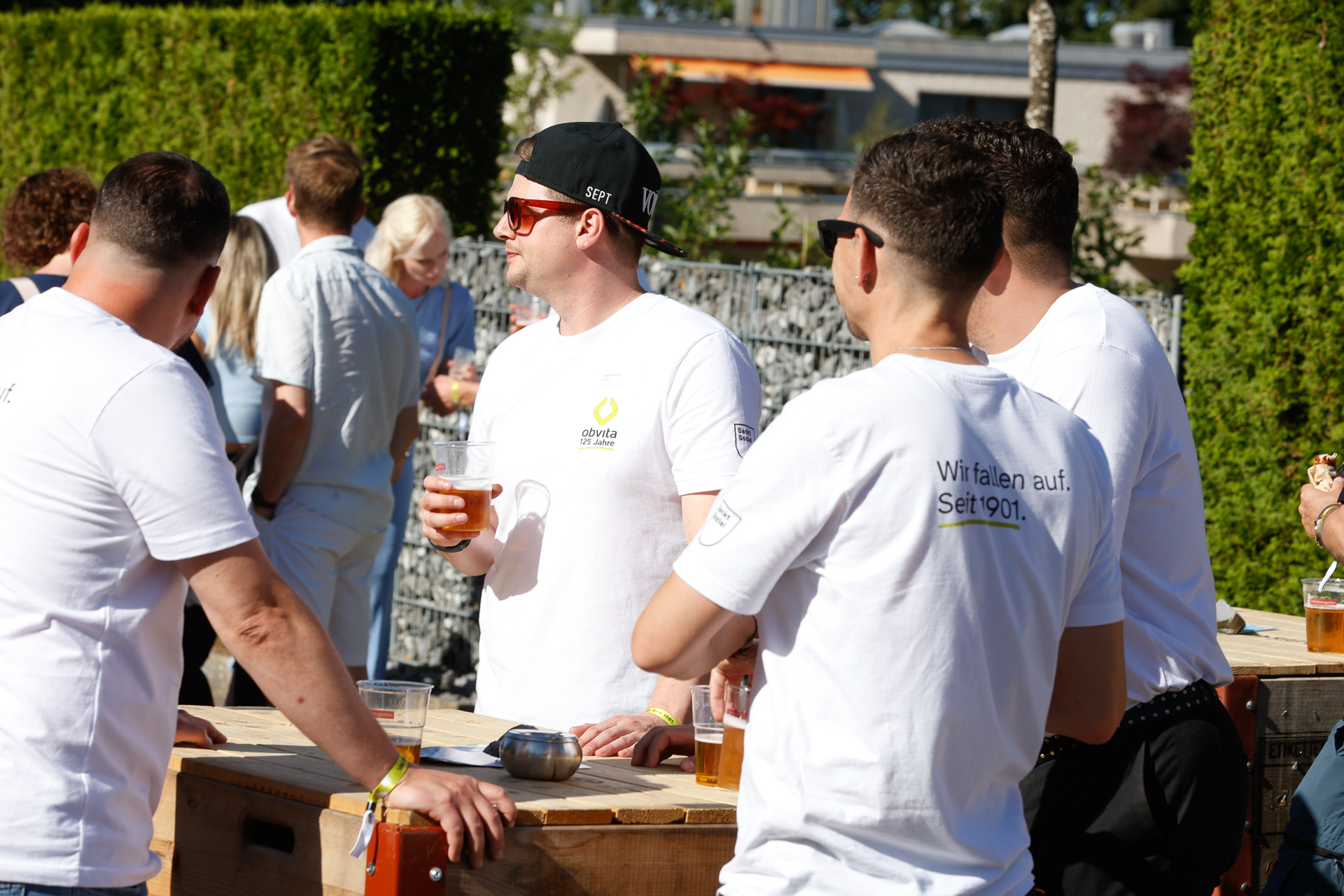 Eine Gruppe von Menschen in weißen T-Shirts steht im Freien um einen Holztisch herum, hält Biergläser in der Hand und unterhält sich im Sonnenlicht. Im Hintergrund sind weitere Personen und Grünpflanzen zu sehen.