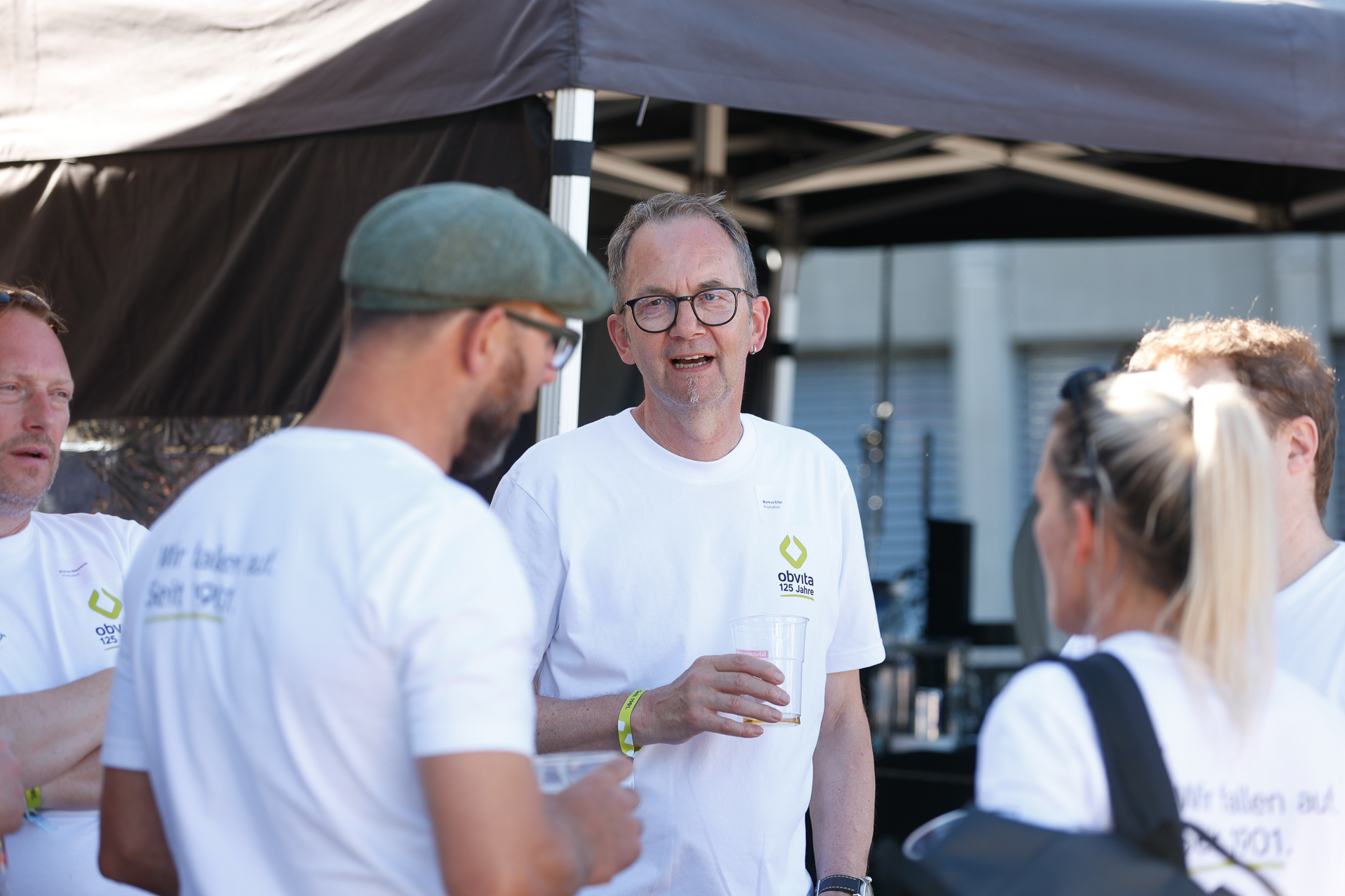 Eine Gruppe von Menschen in weißen T-Shirts steht gemeinsam im Freien, unterhält sich und hält Getränke in den Händen. Ein Mann mit Brille spricht und lächelt; im Hintergrund sind Zelte und Ausrüstung zu sehen.