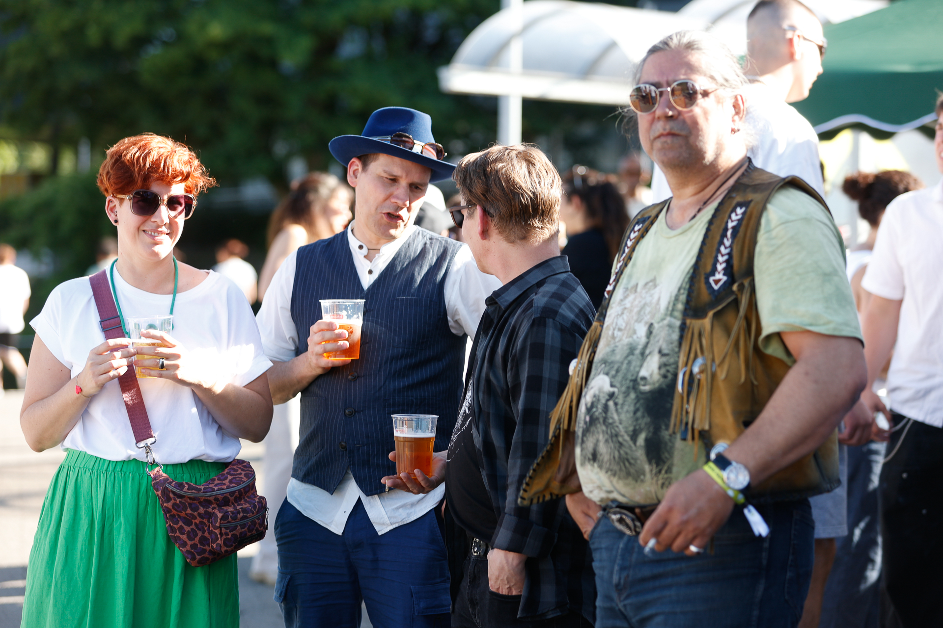 Eine Gruppe von Menschen steht bei einer Veranstaltung im Freien in der Sonne, hält Getränke in der Hand und unterhält sich. Sie tragen legere, farbenfrohe Kleidung und Sonnenbrillen. Im Hintergrund sind Bäume und weitere Teilnehmer zu sehen.