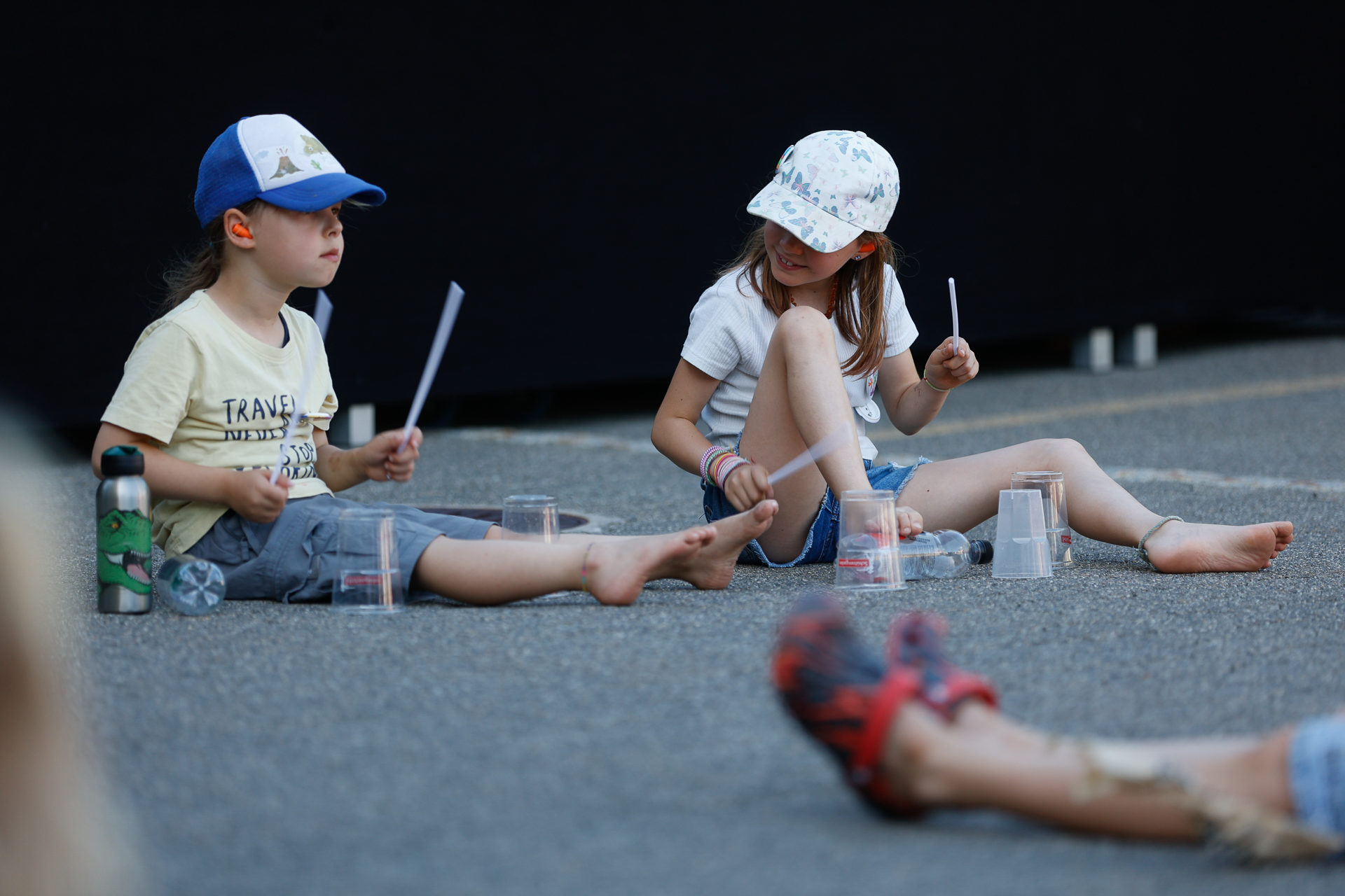 Zwei kleine Kinder sitzen auf dem Boden und machen mit Plastikbechern und Trommelstöcken Musik. Beide tragen Shorts, T-Shirts und Mützen und scheinen ganz in ihre Tätigkeit vertieft zu sein. Die Szene spielt sich im Freien auf einem gepflasterten Untergrund ab.