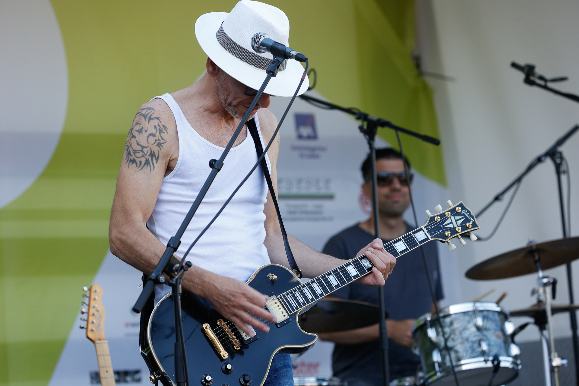Ein Mann mit weißem Hut und Tanktop spielt auf der Bühne E-Gitarre; auf seinem Arm ist ein Tattoo zu sehen. Im Hintergrund ist ein Schlagzeuger zu sehen, der spielt. Die Szene scheint bei einem Live-Musik-Event im Freien zu spielen.
