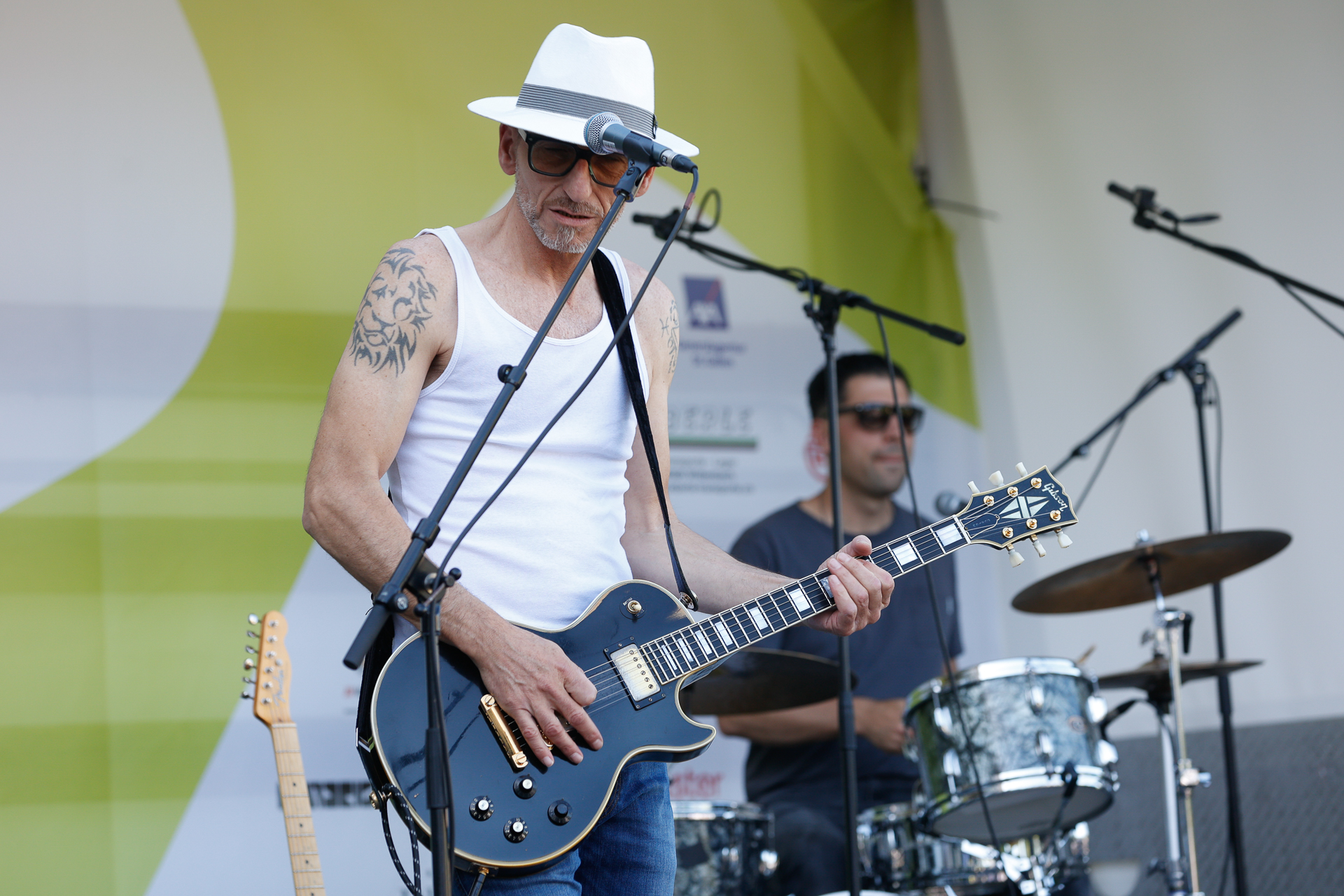 Ein Musiker mit weißem Hut, Sonnenbrille und Trägershirt spielt auf der Bühne E-Gitarre und singt ins Mikrofon, während im Hintergrund ein Schlagzeuger spielt.