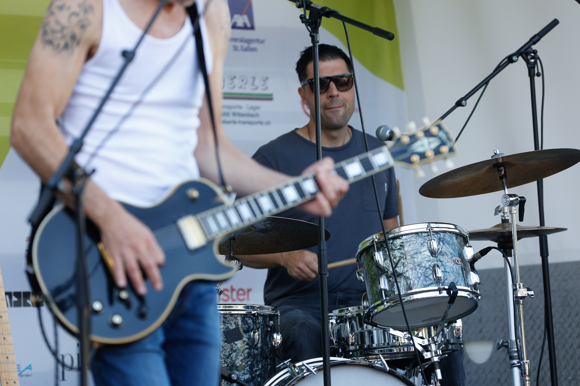 Im Vordergrund spielt ein Musiker auf einer E-Gitarre, während im Hintergrund ein Schlagzeuger mit Sonnenbrille bei einem Open-Air-Konzert am Schlagzeug sitzt. Beide wirken konzentriert und sind von Mikrofonen umgeben.