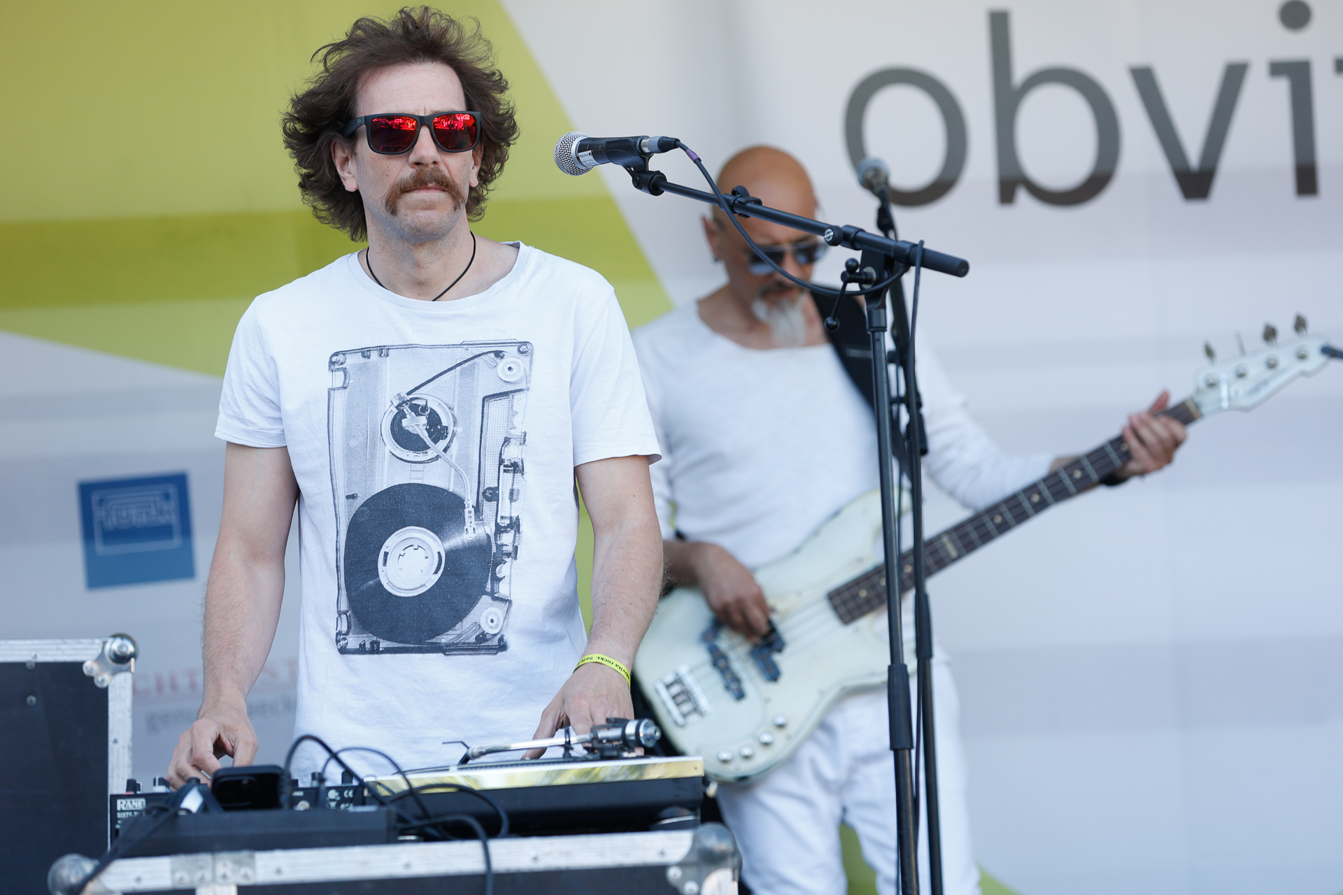 Zwei Musiker treten auf der Bühne auf. Der Musiker im Vordergrund trägt eine Sonnenbrille und ein weißes T-Shirt mit einem Plattenspieler-Motiv, bedient ein DJ-Pult und steht am Mikrofon. Im Hintergrund spielt ein Gitarrist in weißer Kleidung.