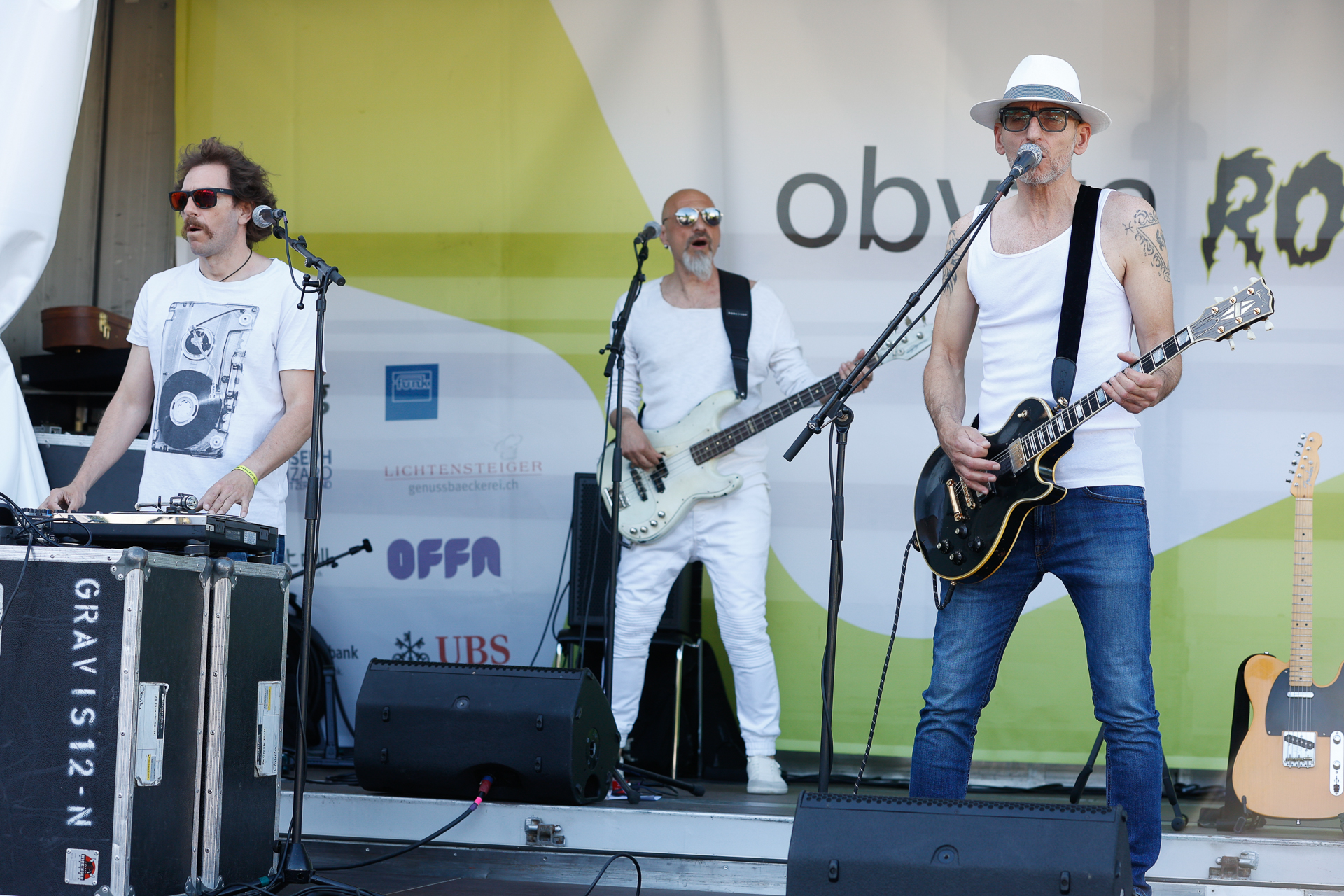 Drei männliche Musiker treten auf einer Freiluftbühne auf. Einer spielt Keyboard, ein anderer Bass, und der dritte, der einen weißen Hut und eine Sonnenbrille trägt, singt und spielt E-Gitarre. Alle tragen weiße Oberteile. Im Hintergrund ist ein Transparent zu sehen.
