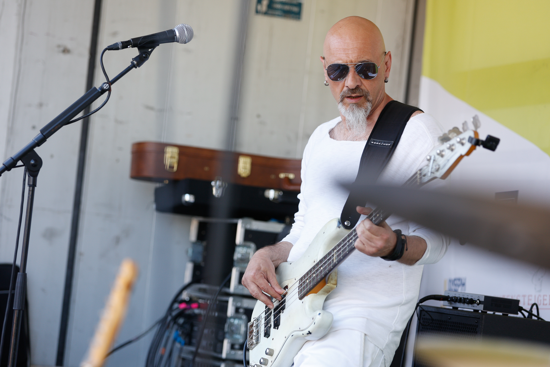 Ein kahlköpfiger Mann mit Bart und Sonnenbrille spielt auf der Bühne E-Bass. Er trägt ein weißes Langarmhemd und eine Hose. Im Hintergrund sind ein Mikrofon, Equipment und ein Gitarrenkoffer zu sehen.