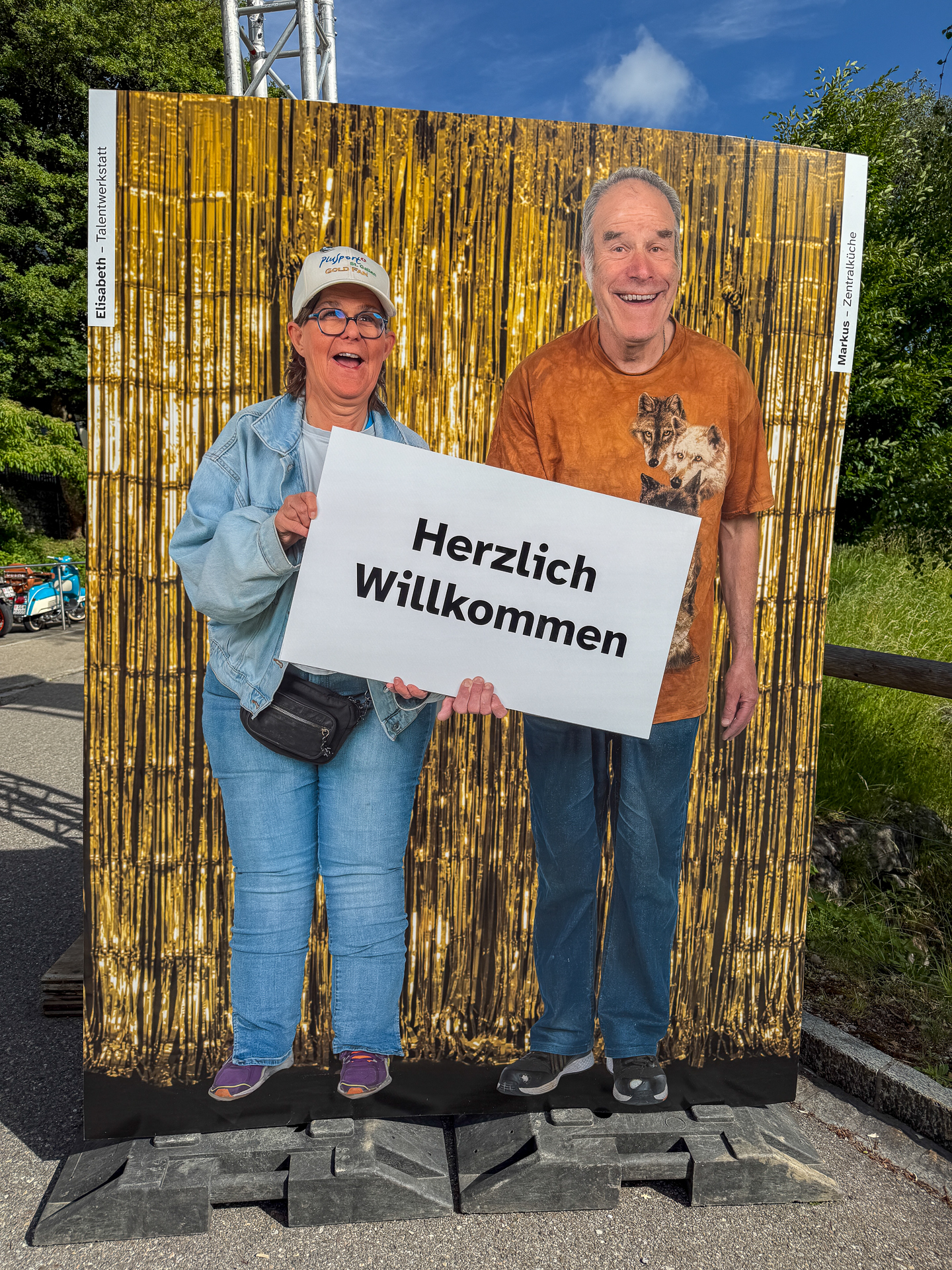 Zwei Personen stehen hinter einer Fototafel mit Ausschnitten und posieren so, als hätten sie Comic-Figuren-Körper. Vor einem glänzenden goldenen Hintergrund halten sie ein Schild mit der Aufschrift „Herzlich Willkommen“ (deutsch für „Herzliches Willkommen“) in die Kamera.