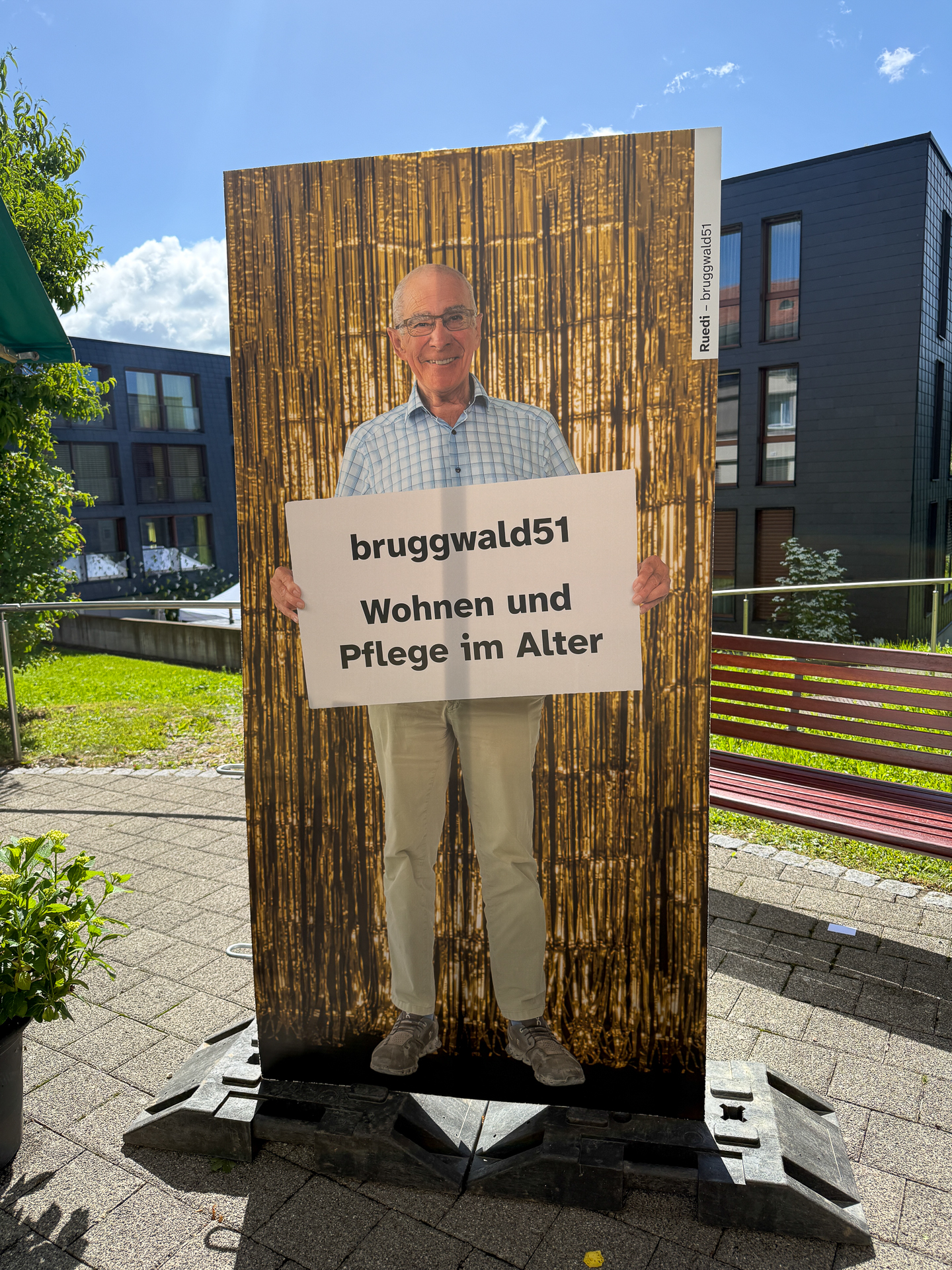 Ein lächelnder Mann hält ein Schild mit der Aufschrift „bruggwald51 Wohnen und Pflege im Alter“ in der Hand, während er im Freien vor einem goldfarbenen Paillettenhintergrund steht; im Hintergrund sind moderne Gebäude und Grünflächen zu sehen.