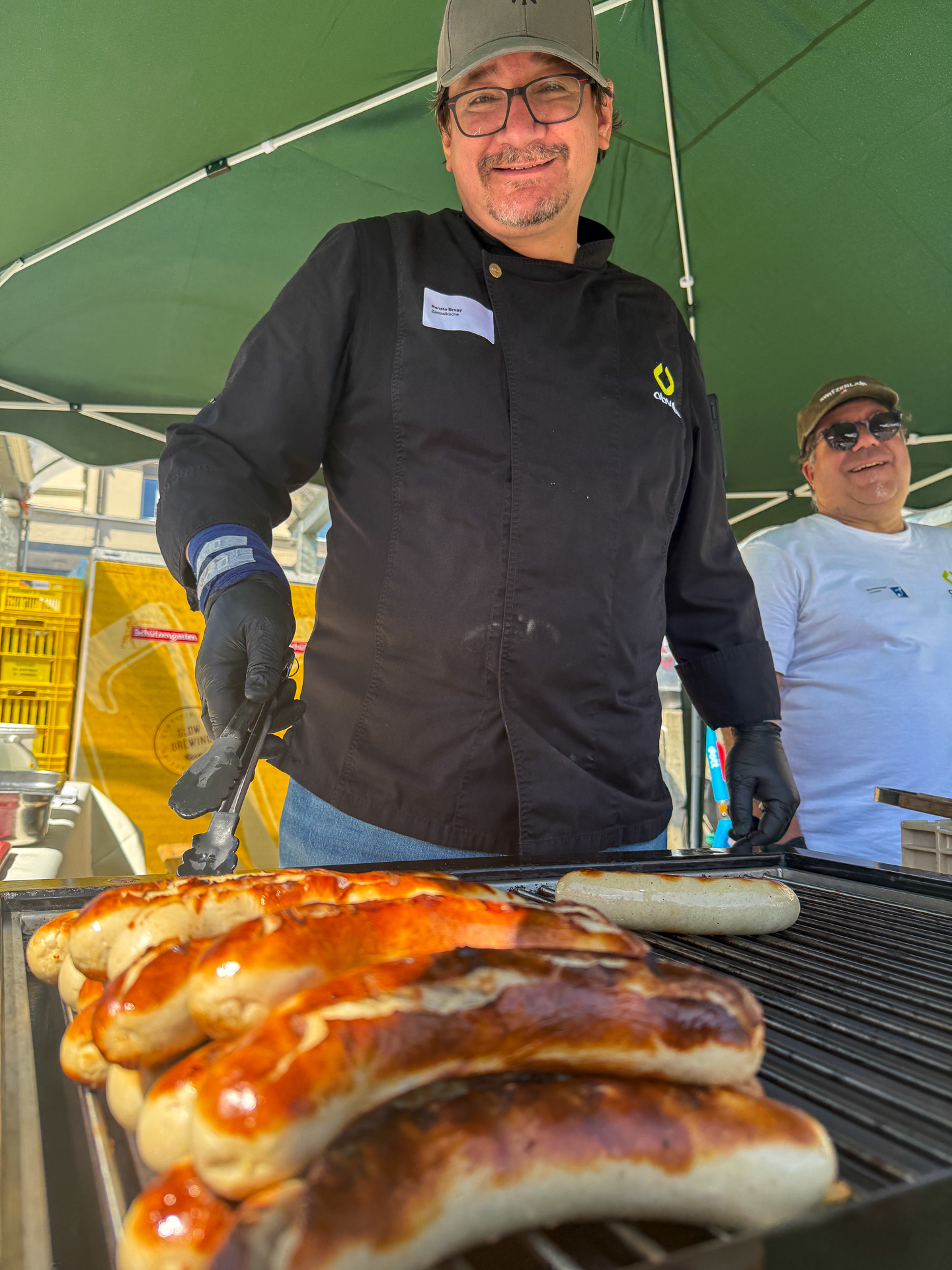 Ein lächelnder Mann in schwarzer Kochjacke und Kochmütze grillt draußen unter einem grünen Vordach Würstchen. Im Hintergrund steht ein weiterer Mann mit Sonnenbrille und weißem Hemd, der ebenfalls lächelt.