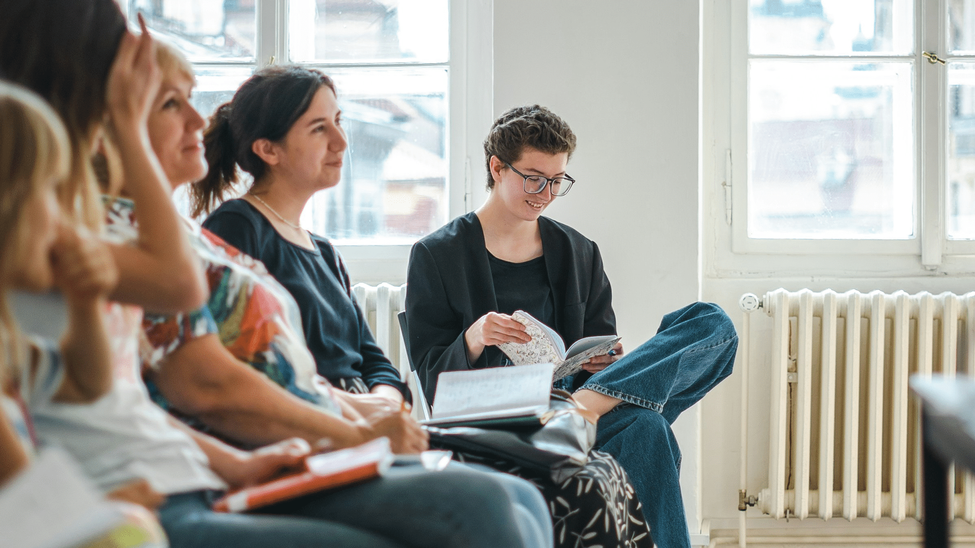 In einem hellen Raum mit großen Fenstern sitzen mehrere Personen, von denen einige Notizbücher in der Hand halten. Eine Person sitzt lächelnd etwas abseits und liest ein Buch; sie wirkt konzentriert und entspannt.