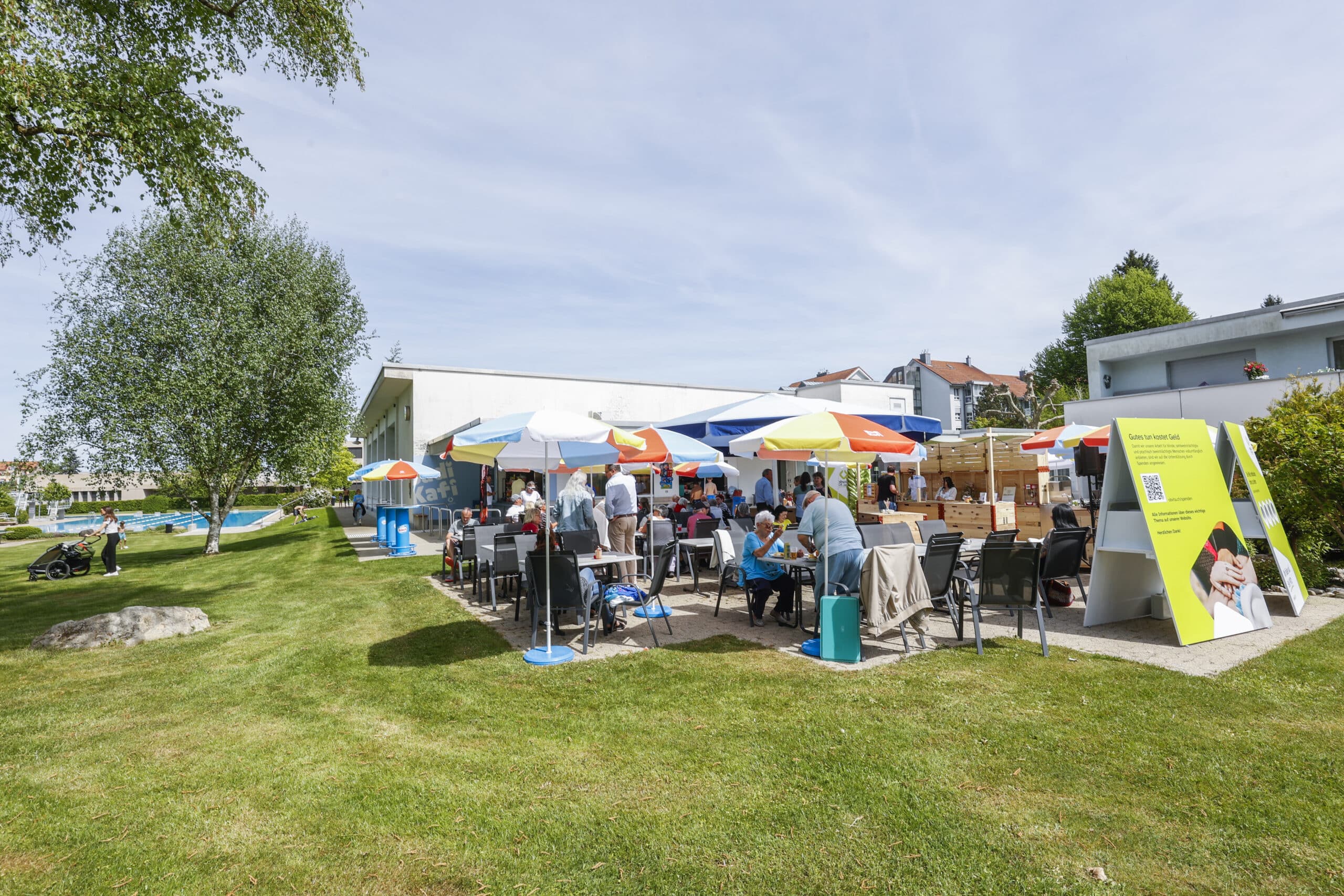 An einem sonnigen Tag versammeln sich Menschen an Tischen im Freien unter bunten Sonnenschirmen in der Nähe eines modernen weißen Gebäudes. Im Vordergrund sind ein großes Schild und grünes Gras zu sehen. Im Hintergrund sind Bäume und weitere Gebäude zu erkennen.