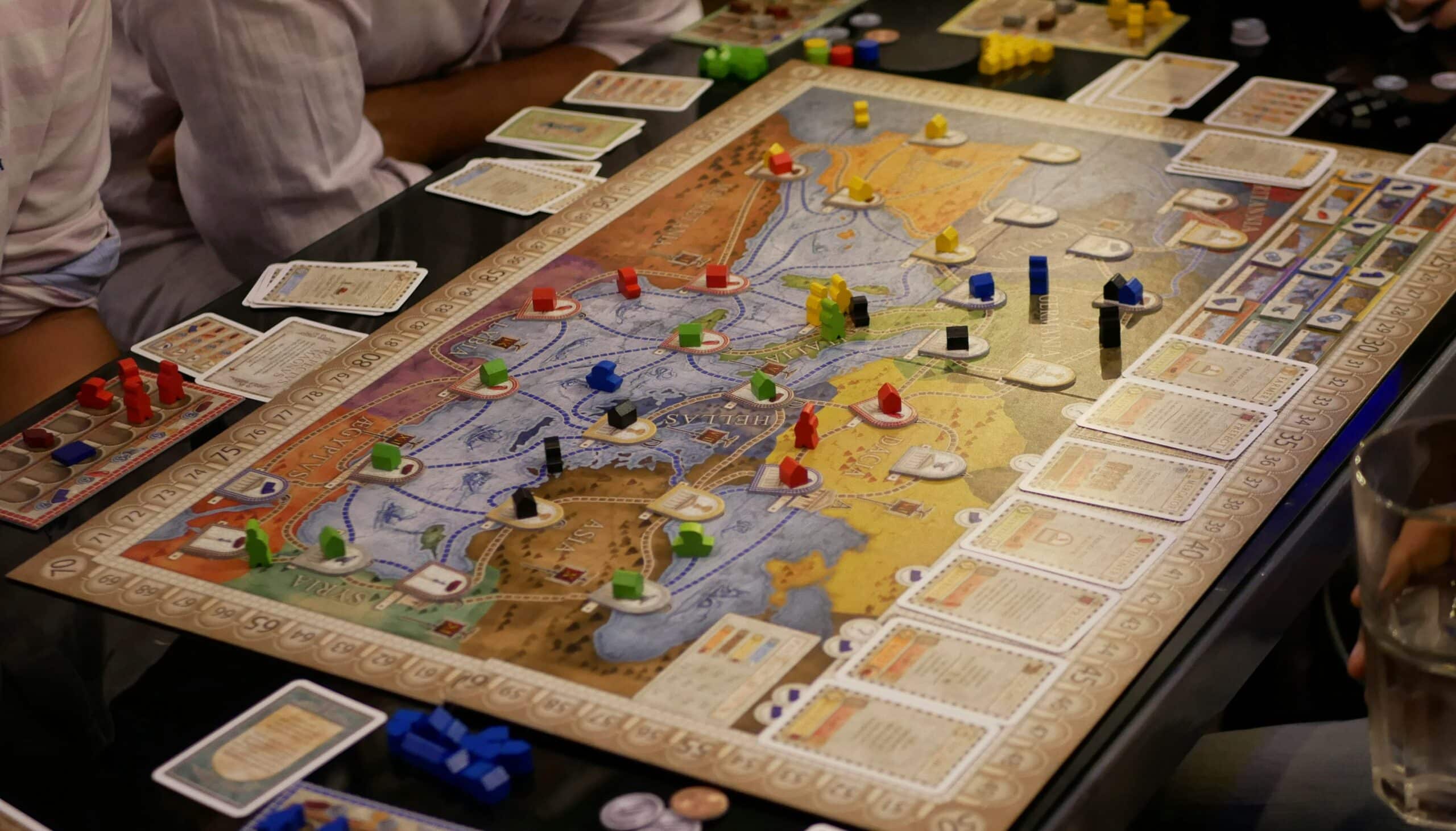 Vier Personen spielen ein strategisches Brettspiel mit bunten Spielfiguren, Karten und einer detaillierten Karte. Die Spielkomponenten sind auf einem schwarzen Tisch ausgebreitet, und in der Ecke steht ein Glas Wasser.